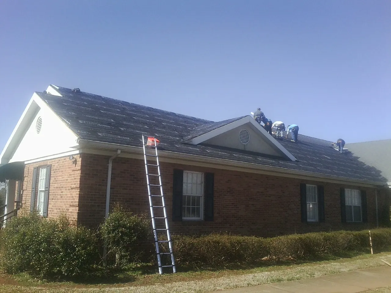 Crew tearing shingles off a brick ranch roof with a ladder set — Metal Roof Repair work by our crew in Osterville