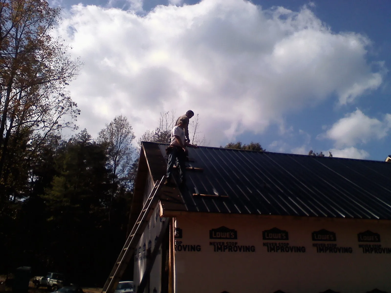Roofer fastening dark metal panels high against a cloudy sky, from a Roof Inspection job in Osterville
